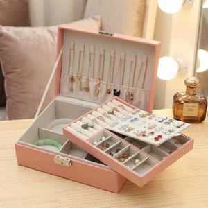Puede incluir: Una caja de joyería rosa con múltiples compartimentos y una tapa con bisagras. La caja está abierta, revelando varios collares, anillos y pendientes. El interior está forrado con una tela de color claro, y las joyas se exhiben cuidadosamente. Un frasco de perfume está en el fondo.
