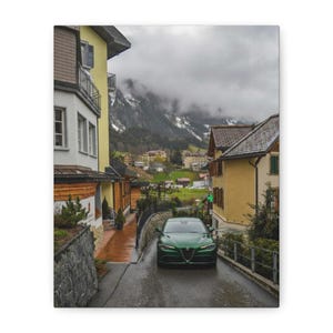 May include: A green sports car drives down a wet road in a European village. The car is the focal point, with buildings and mountains in the background. The sky is overcast, and the scene has a muted color palette.