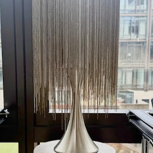 May include: A table lamp featuring a cylindrical shade constructed from numerous hanging silver-toned metal chains. The lamp has a smooth, flared, brushed silver-toned metal base that supports the chain fringe, creating a cascading effect.