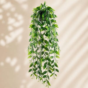 May include: A hanging artificial plant featuring long, cascading stems with pointed, variegated green leaves. The foliage displays a mix of light and dark green shades, extending downward from a simple, dark-colored circular pot at the top.