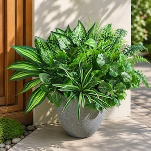 May include: A large, rounded, speckled gray planter filled with a dense arrangement of various artificial green plants, including broad leaves with light green stripes, delicate fern fronds, and rounded foliage, creating a lush, full display.