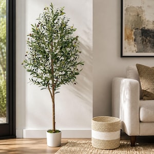 May include: A tall artificial olive tree in a white pot. The tree has green leaves and small olive-like fruits. The trunk is brown, and the pot is cylindrical. The tree is placed on a woven rug near a woven basket.