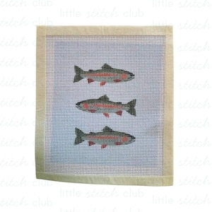 May include: A square needlepoint canvas featuring a light blue background with three horizontally aligned fish. Each fish is rendered in shades of gray, white, and red, with a visible grid pattern throughout the design and a plain beige border.
