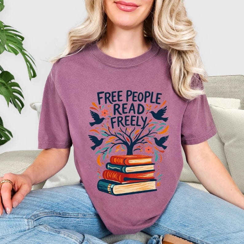 May include: A mauve-colored t-shirt with the text "FREE PEOPLE READ FREELY" in orange and blue. The design includes a tree, birds, and a stack of books. The shirt is a comfortable, casual style.