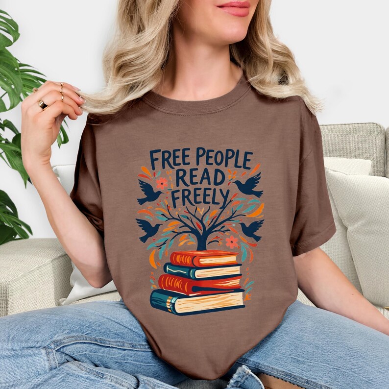 May include: Brown t-shirt with the text "FREE PEOPLE READ FREELY" in blue. The design includes a tree, birds, and books in orange, blue, and green. The shirt is a casual, short-sleeved style.