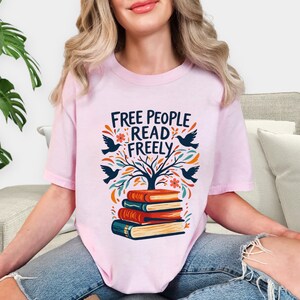 May include: A light pink t-shirt with the text "FREE PEOPLE READ FREELY" above a graphic of a tree, birds, flowers, and a stack of books. The design is in shades of blue, orange, and green.