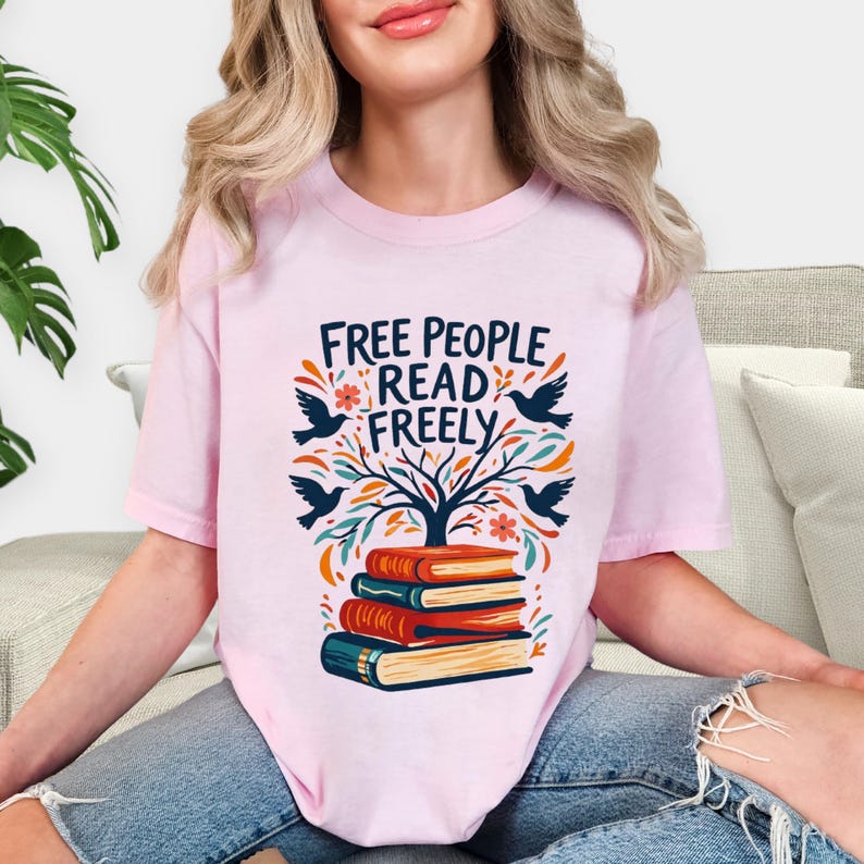 May include: A light pink t-shirt with the text "FREE PEOPLE READ FREELY" above a graphic of a tree, birds, flowers, and a stack of books. The design is in shades of blue, orange, and green.