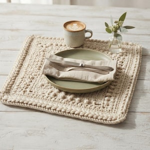 Puede incluir: Un mantel individual cuadrado de ganchillo color crema con un patrón texturizado de puntos de burbuja y un borde trenzado. Centrado sobre el mantel hay un plato verde salvia, una servilleta de lino beige doblada, un tenedor y un cuchillo de plata.