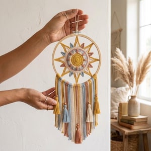 Könnte beinhalten: Ein runder Wandbehang aus einem Holzring mit einem gehäkelten Sonnenmotiv in Gelb, Blau und Creme. Unter der Sonne hängen mehrere Garnstränge in Blau, Creme und Terrakotta, die mit kleinen Holzperlen und dekorativen Quasten verziert sind.