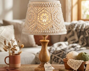 May include: A table lamp with a light wood base and a cream-colored crocheted lampshade featuring intricate geometric patterns. A small white tag attached to the base reads THE LUMIMA LAMP, Handmade Crochet, Pattern by ArtisanCrafts, EngridDesign, Pattern Included.