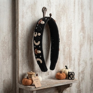 Könnte beinhalten: Ein schwarzes Häkel-Stirnband mit gestickten cremefarbenen und braunen Pilzmotiven, Mondsichelformen und kleinen weißen Kreuzstich-Akzenten. Das Stirnband hängt an einem Metallhaken, daneben befinden sich kleine gehäkelte Kürbisse.
