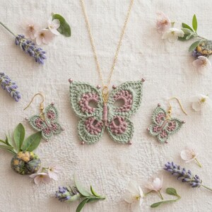 May include: A jewelry set consisting of a necklace and a pair of earrings, each featuring a crocheted butterfly motif in sage green and dusty rose. The butterfly pendant hangs from a thin gold-tone chain, while the earrings have gold-tone hooks.
