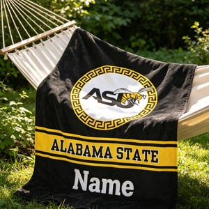 Op de afbeelding: Zwarte strandlaken met gele strepen en de witte woorden "ALABAMA STATE". De handdoek heeft een cirkelvormig logo met "ASU" en een gele horzel. Het woord "Name" is ook zichtbaar.