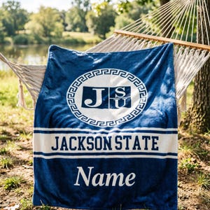 Puede incluir: Una manta azul de felpa con un emblema circular blanco con las letras JSU, una franja horizontal blanca con el texto JACKSON STATE en azul oscuro y la palabra Name impresa en blanco en la parte inferior, sobre una textura de tela suave.