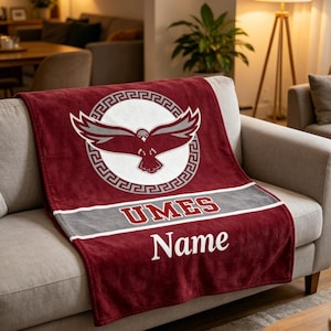 Puede incluir: Una manta granate con una franja blanca y gris, con un logotipo de un pájaro con las alas extendidas dentro de un diseño circular. Los textos "UMES" y "Name" se muestran en blanco.