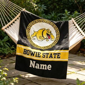 Puede incluir: Una manta rectangular negra con un emblema circular amarillo y blanco que muestra una ilustración de un bulldog. Debajo del emblema, una franja horizontal amarilla contiene el texto BOWIE STATE en negro, seguido de la palabra Name en blanco.