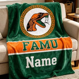 Könnte beinhalten: Grün-orange FAMU-Decke mit Schlangenlogo in einem Kreis. Der Schriftzug "FAMU" ist orange und weiß, und das Wort "Name" ist weiß. Die Decke liegt über einem beigen Sofa.