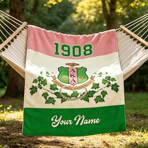Op de afbeelding: Een roze, witte en groene deken met de tekst "1908" in groen. De deken heeft een wapen met klimop en de woorden "Your Name" in witte letters. De deken is over een hangmat gedrapeerd.
