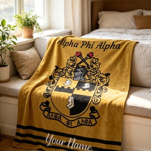 May include: Mustard yellow blanket with the words "Alpha Phi Alpha" and a crest design. The crest features a knight's helmet, crossed torches, and the Greek letters Alpha, Phi, and Alpha. The bottom of the blanket has black stripes and the words "Your Name".