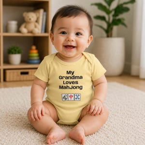 May include: A light yellow baby bodysuit featuring the black text 'My Grandma Loves MahJong' followed by three Mahjong tile illustrations, including a bird, a red character, and a five-dot tile, printed on the front of the garment.