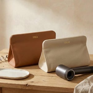 May include: Two leather cosmetic bags, one tan and one light beige, sit on a wooden surface. The tan bag is embossed with "TINA", and the beige bag has "F.G.H". A hair dryer and a small white plate are also on the table.