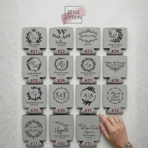 May include: A collection of sixteen light gray foam can coolers arranged in a four-by-four grid. Each cooler features a unique black printed design, including monograms, floral wreaths, and names with dates, labeled with numbers from 21 to 32.