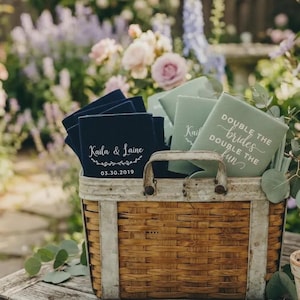 May include: A woven wooden basket filled with navy blue and sage green foam can coolers. The navy coolers feature the text Kaila & Laine with a date of 03.30.2019, while the sage green coolers display the text Double the brides, double the fun.