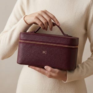 以下が含まれることがあります： ゴールドのジッパーとハンドルが付いた、バーガンディ色の長方形の化粧ケース。ケースには金色のイニシャル「BG」が入っています。クリーム色のセーターを着た人がケースを持っています。