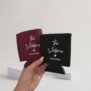 May include: Two foam can coolers, one burgundy and one black, each featuring white printed text that reads The Watsons, a small heart icon, and the date 10.22.2022. The sleeves are designed to hold beverage cans and have a flat base.