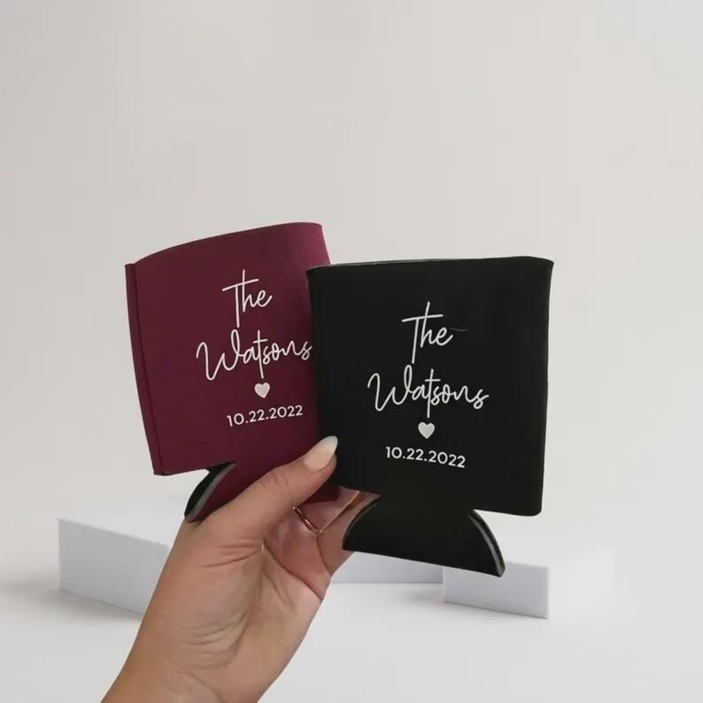 May include: Two foam can coolers, one burgundy and one black, each featuring white printed text that reads The Watsons, a small heart icon, and the date 10.22.2022. The sleeves are designed to hold beverage cans and have a flat base.