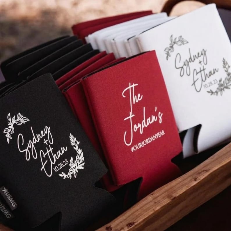 May include: A collection of foam can coolers in black, red, and white. The black and white coolers feature a floral wreath design with the text Sydney + Ethan 10.28.23. The red cooler displays the text The Jordan's and the hashtag #OURJORDANYEAR.