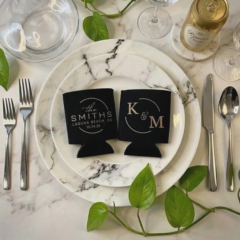 May include: Two black foam can coolers featuring white printed text. One cooler displays the text the SMITHS LAGUNA BEACH, CA 10.24.26, and the other displays the letters K & M, both enclosed within a circular graphic design.