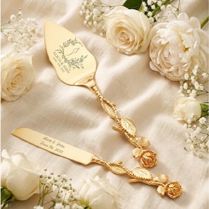 May include: Gold-colored cake server and knife set with floral and leaf detailing. The server and knife are engraved with the names "Alicia & John" and the date "June 7th, 2023". The handles feature rose and vine designs.