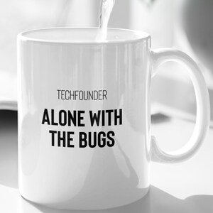 Peut inclure: Une tasse en céramique blanche avec le texte "TECHFOUNDER ALONE WITH THE BUGS" en noir. De l'eau est versée dans la tasse depuis une cruche foncée. Une plante verte est en arrière-plan.