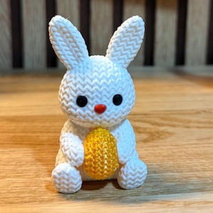 Pode incluir: Uma figura de coelho branco de malha com olhos pretos e nariz vermelho, segurando um ovo amarelo. O coelho tem orelhas compridas e está sentado em posição vertical. A figura tem aproximadamente 7,6 cm de altura.
