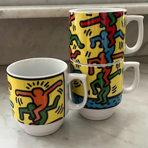 Pode incluir: Conjunto de três canecas de cerâmica branca com fundo amarelo e um design abstrato e colorido de figuras dançando. Cada caneca tem uma alça branca e um interior branco. As canecas são empilhadas e dispostas em uma superfície de mármore.