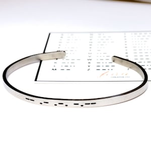 May include: A silver-colored cuff bracelet with Morse code etched on the front. A card with a Morse code alphabet chart is in the background. The bracelet is open-ended and sits on a white surface.