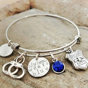 Puede incluir: Pulsera de brazalete plateada con m&uacute;ltiples dijes. Los dijes incluyen esposas, un dije redondo con el texto "My hero wears blue", una gema azul y una placa de polic&iacute;a. La pulsera se muestra sobre una superficie texturizada.