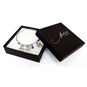 Puede incluir: Un brazalete plateado con m&uacute;ltiples dijes, incluyendo una gema azul, una insignia y un dije que dice "My hero wears blue". El brazalete se exhibe en una caja de regalo negra abierta con las palabras "fierce creative co." en letras doradas.
