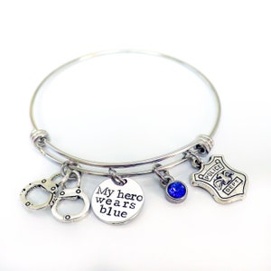 Puede incluir: Pulsera de brazalete plateada con m&uacute;ltiples dijes. Los dijes incluyen esposas, un dije redondo con el texto "My hero wears blue", una gema azul y una placa de polic&iacute;a. Fondo blanco.