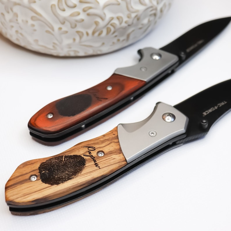 May include: Two pocket knives with wooden handles. One handle is brown wood with a black fingerprint design and the text "Papa" engraved on it. The other handle is light brown wood with a black fingerprint design and the text "Grandpa" engraved on it.