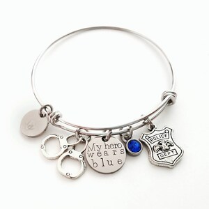 Puede incluir: Pulsera de brazalete plateada con m&uacute;ltiples dijes. Incluye esposas, un dije redondo con el texto "My hero wears blue", una gema azul y una placa de polic&iacute;a. Sobre un fondo blanco.