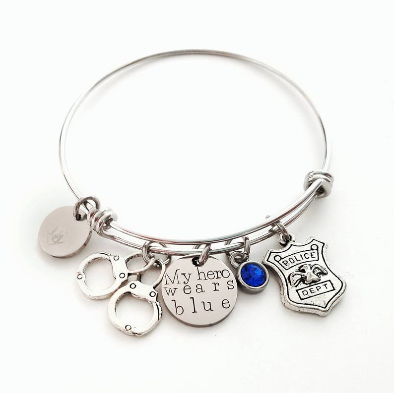Puede incluir: Pulsera de brazalete plateada con m&uacute;ltiples dijes. Incluye esposas, un dije redondo con el texto "My hero wears blue", una gema azul y una placa de polic&iacute;a. Sobre un fondo blanco.