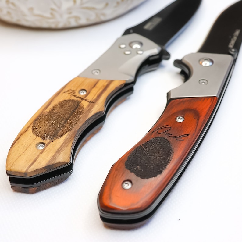 May include: Two pocket knives with wooden handles. One handle is light brown with the word "Mom" engraved on it. The other handle is dark red with the word "Dad" engraved on it.
