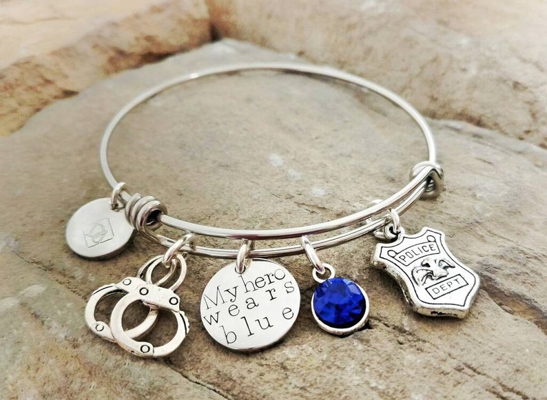 Puede incluir: Pulsera de brazalete plateada con m&uacute;ltiples dijes. Los dijes incluyen esposas, un dije redondo con el texto "My hero wears blue", una gema azul y una placa de polic&iacute;a. La pulsera se muestra sobre una superficie texturizada.