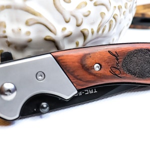 May include: A brown wooden pocket knife with a silver handle. The knife has a personalized inscription that reads "Paul" and a fingerprint design. The knife is labeled "TRC-F5".