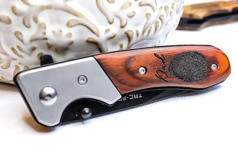 May include: A brown wooden pocket knife with a silver handle. The knife has a personalized inscription that reads "Paul" and a fingerprint design. The knife is labeled "TRC-F5".