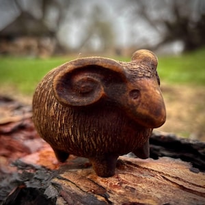Puede incluir: Una pequeña figura de carnero de madera tallada intrincadamente. El carnero es de color marrón cálido con texturas detalladas, incluyendo cuernos rizados y un pelaje texturizado. La figura tiene cuatro patas pequeñas.