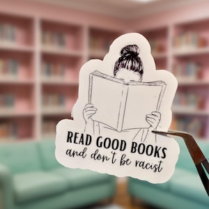 May include: A white sticker featuring a line drawing of a person with their hair in a bun, reading a book. The text "READ GOOD BOOKS and don't be racist" is below the illustration. The sticker is held by tweezers.