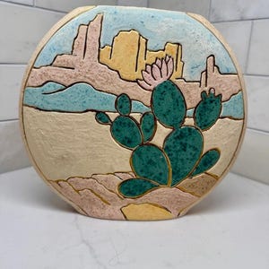 May include: A round ceramic vase featuring a raised relief design of a desert landscape. The scene depicts a green prickly pear cactus with a pink flower in the foreground, set against a background of tan mesas, blue sky, and textured sandy ground.